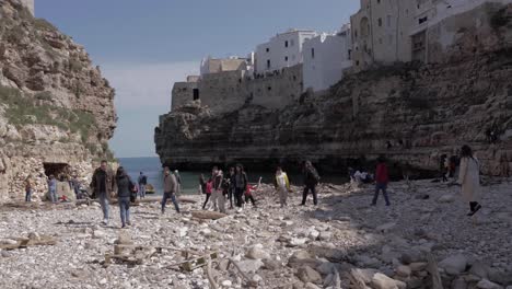 People-walking-down-a-rocky-beach-in-Polignano-a-Mare,-enjoying-wonderful-nature-on-the-seaside-shore