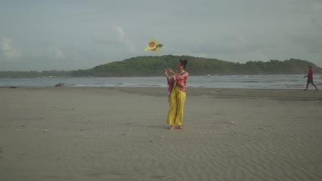 Ein-Junges-Asiatisches-Model-In-Gelbem-Outfit-Lässt-Ihren-Drachen-Freudig-Fallen,-Während-Sie-An-Einem-Strandort-Steht