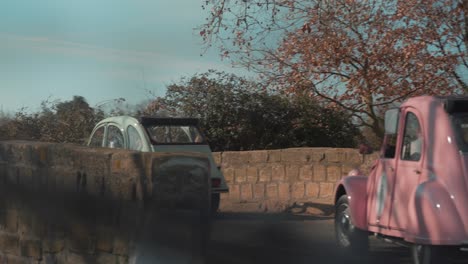 Three-vintage-cars-are-driving-along-a-small-road,-passing-over-a-stone-bridge