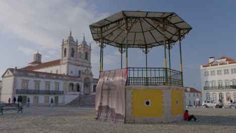 Die-Kirche-Hinter-Der-Nazare-Beschilderung-Mit-Touristen,-Die-Durch-Das-Stadtzentrum-Am-Meer-In-Portugal-Spazieren