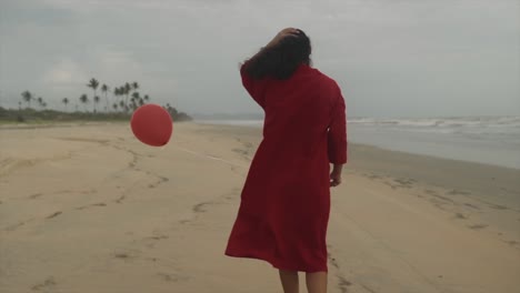 A-an-exquisite-Asian-model,-elegantly-adorned-in-a-crimson-long-top,-gripping-a-scarlet-balloon-as-she-gazes-towards-the-seashore-while-gracefully-strolling