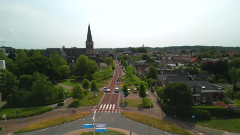 Drone-shot-of-giant-roundabout-in-Castricum,-the-Netherlands,-with-flowing-traffic,-surrounded-by-trees-and-buildings,-daytime-during-summer