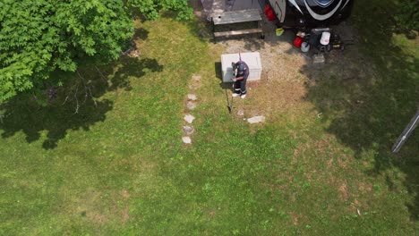 View-From-Above-Of-Male-Camper-Trimming-Grass-With-Brush-Cutter-At-The-Campsite