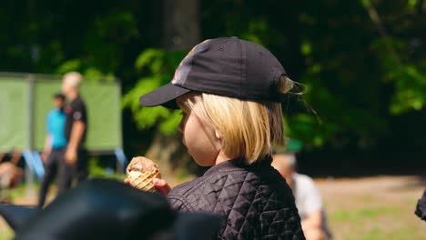 Capture-the-pure-joy-of-a-7-year-old-blonde-boy-with-long-hair,-wearing-a-cap,-savoring-an-ice-cream-cone-in-a-sunny-park-or-forest