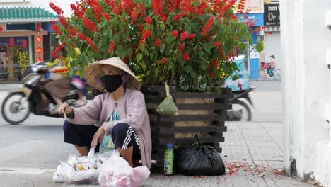 Maskierter-Straßenverkäufer-Sitzt-An-Einer-Belebten-Straße-Neben-Blumen