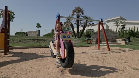 Little-Girl-Riding-a-Toy-Motorcycle-in-the-Sandbox-at-Urban-Park,-Hammock-and-Slide,-Outdoors-Playground-at-Calders-Spain-Barcelona