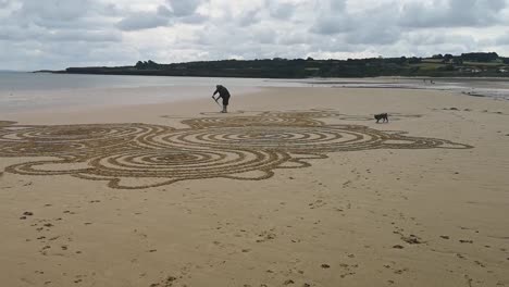 Male-creating-artistic-flowing-patterns-on-sandy-Anglesey-beach-with-his-terrier-dog