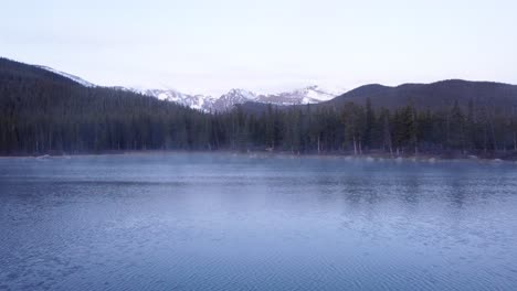 Steam-rising-off-of-Colorado-mountain-lake-with-snowcapped-Rocky-Mountains-in-the-background,-pan-left-aerial