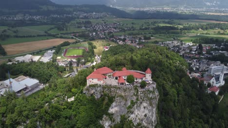 Luftaufnahme-Des-Berühmten-Burgbergs-In-Bled,-Umgeben-Von-Bauernhöfen-Und-Fußballfeldern-In-Einer-Berglandschaft-Bei-Grauen-Wolken-Am-Himmel