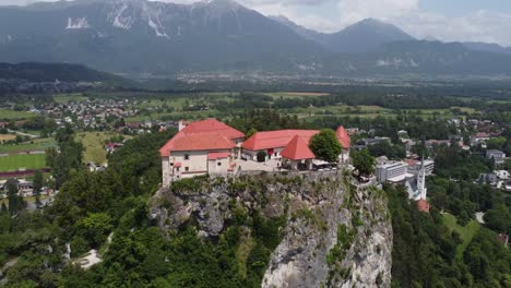 Aerial-backwards-flight-showing-old-castle-in-bled-located-on-hill-during-sunny-day,-Slovenia---rising-flight
