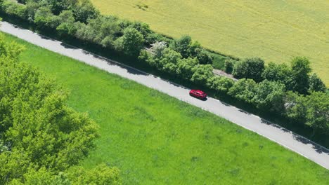Tracking-Drone-Shot-of-Red-Car-Moving-on-Countryside-Road-on-Sunny-Summer-Day,-Between-Meadows,-Trees-and-Railway