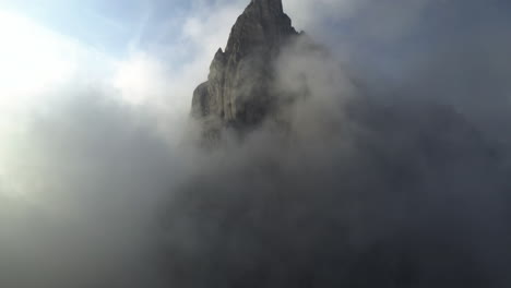 Drone-shot-tilting-toward-a-rocky-peak-surrounded-by-low-hanging-clouds-in-Italy