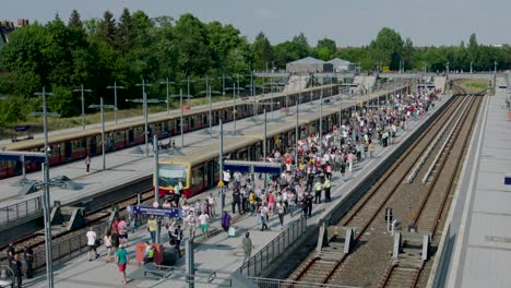 An-outdoor-scene-depicting-a-bustling-train-station-with-multiple-tracks-and-platforms,-featuring-a-crowd-of-people,-trains,-and-encircled-by-trees-and-buildings