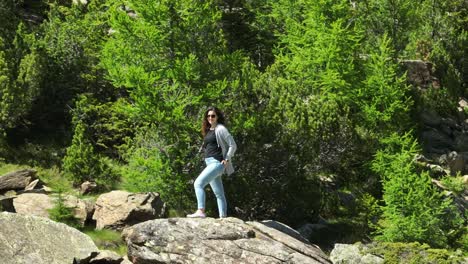 Brunette-woman-with-sunglasses-poses-standing-on-rock-surrounded-by-nature-of-Val-Masino-in-Valtellina,-Italy