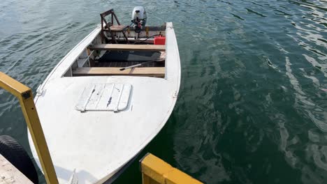 Small-white-fishing-motor-boat-with-pointed-bow-floating-on-top-of-a-calm-lake,-tied-at-the-port-on-a-sunny-summer-day