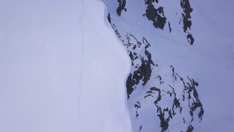 Vista-Aérea-De-La-Cresta-A-Lo-Largo-De-Las-Montañas-Parang-Cubiertas-De-Nieve-Con-Huellas-De-Escalador-Y-Pendiente-Empinada-Con-Rocas-Dentadas