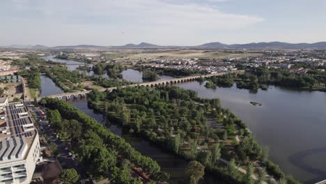 Aerial-view-orbiting-riverbank-regeneration-on-Guadina-river-with-the-roman-bridge-of-Merida-crossing-scenic-blue-water