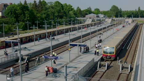 Bahnhof-Im-Freien-Voller-Menschen-Auf-Bahnsteigen,-Mehrere-Züge-Auf-Gleisen-Unter-Blauem-Himmel,-Eingerahmt-Von-Bäumen-Und-Gebäuden