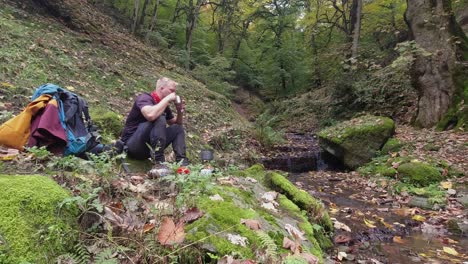 Young-male-hiker-enjoys-hot-tea-and-snack-in-forest-beside-tiny-stream