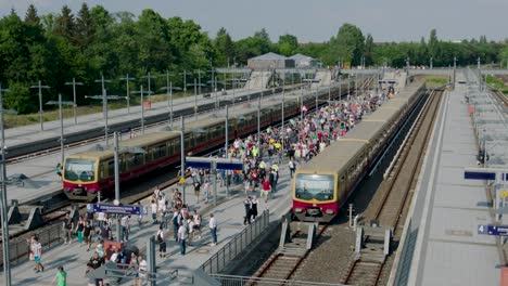 Camera-tilt-capturing-outdoor-train-station,-crowded-platforms,-trains,-signs,-lampposts-under-blue-sky,-surrounded-by-trees-and-buildings