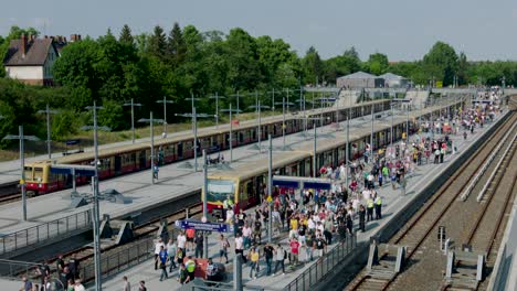 Camera-tilt-captures-a-vibrant-outdoor-train-station-with-multiple-platforms,-trains,-bustling-crowds,-signs,-lampposts,-trees-and-buildings-under-a-blue-sky