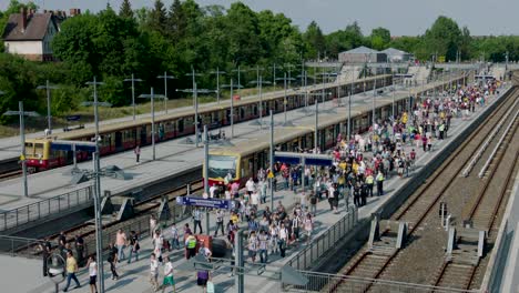 Outdoor-train-station-bustling-with-people,-trains-on-tracks-under-a-blue-sky,-surrounded-by-trees-and-buildings,-captured-by-a-static-camera