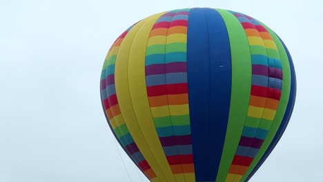 Descending-hot-air-balloon-on-overcast-day,-aerial-close-up-of-multi-colored-balloon-with-flame-of-fire,-people-in-wicker-basket