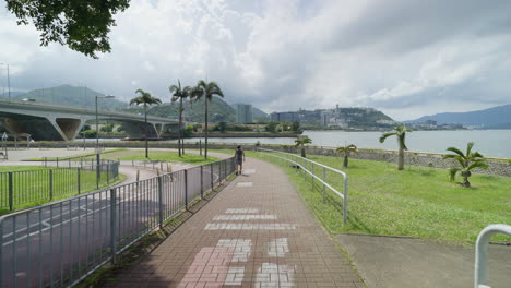 Caminando-Por-Un-Camino-De-Ladrillo-Sobre-El-Río-Con-La-Autopista-Y-Los-Edificios-De-Hong-Kong-En-La-Distancia