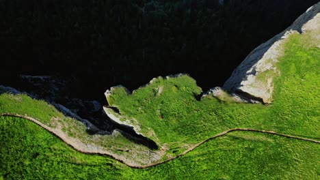 Sheer-cliff-on-side-of-grassy-green-mountaintop-with-trail,-bird’s-eye-view