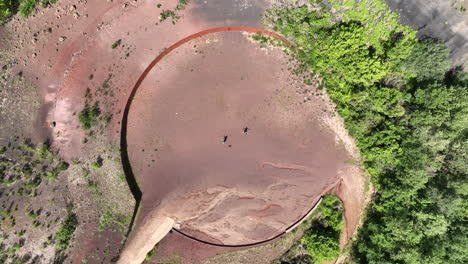 Top-down-drone-view-of-two-people-and-dog-running-inside-unusual-circle-shaped-volcanic-area