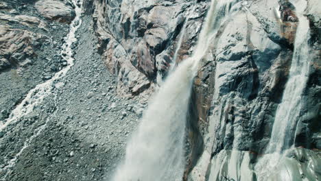 Giant-waterfall-coming-from-a-Glacier-Melting-in-a-lake-below-due-to-Climate-Change-in-the-Alps,-Fellaria-Big-Waterfalls-come-down-from-the-glaciers-edge,-top-down-view-of-the-mountains-and-lake-below