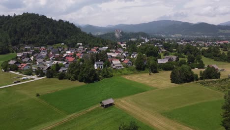 Sobrevuelo-Aéreo-Campos-Agrícolas-Y-Pequeña-Ciudad-De-Bled-Con-Montañas-Y-Clips-En-El-Fondo,-Eslovenia