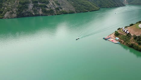 Albania,-a-boat-sailing-through-the-green-waters-of-Lake-Koman-amidst-the-slopes-of-the-Accursed-Mountains,-aerial-view-from-a-drone