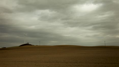 White-wind-turbine-atop-a-hill-forms-the-backdrop-for-a-bleak-and-desolate-vast-field-landscape-dotted-with-telephone-poles-under-an-overcast-sky