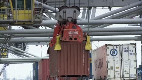 Close-up-cargo-crane-lifts-a-red-ship-container,-loading-vessel-at-harbor