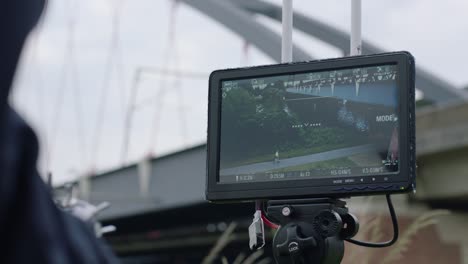 Handheld-camera-view-of-a-monitor-on-tripod-showing-live-feed-from-a-drone-of-white-curved-arch-bridge-under-overcast-sky,-blurred-person-in-foreground