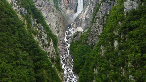 Aerial-forward-flight-over-rocky-river-between-steep-greened-mountain-cliffs-and-splashing-waterfall-in-backdrop---Posocje,-Slovenia