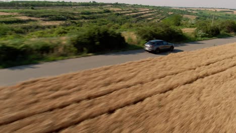 Aerial-tracking-shot,-of-a-grey-luxury-SUV-Sport-car,-driving-on-a-country-tarmac-road,-surrounded-by-golden-wheat-fields