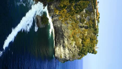 Vertical-Slow-motion-reveal-shot-of-the-cliffs-of-Pura-Uluwatu-temple-in-Bali,-Indonesia-overlooking-the-beautiful-cliffs-and-high-waves-from-the-blue-sea