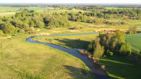 Curved-river-flowing-through-meadows,-aerial-view-in-golden-hour-with-long-tree-shadows