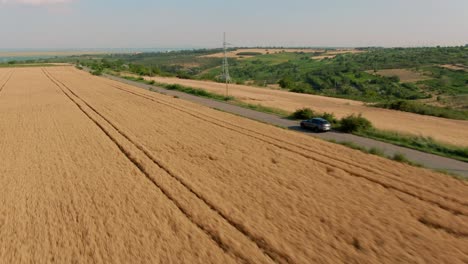 Aerial-tracking-shot-of-a-grey-luxury-SUV-Sport-car-driving-slowly-on-a-country-tarmac-road-surrounded-by-golden-wheat-fields
