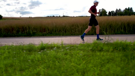A-man-dressed-in-sports-gear,-a-white-hat,-running-shoes,-and-sunglasses-is-running-on-a-road-against-a-backdrop-of-cultivated-fields