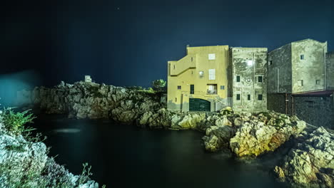 River-timelapse-with-the-buildings-on-the-rocks-on-its-bank-in-the-night-time-with-dark-sky-in-the-background-in-Sicily,-Island-of-Italy