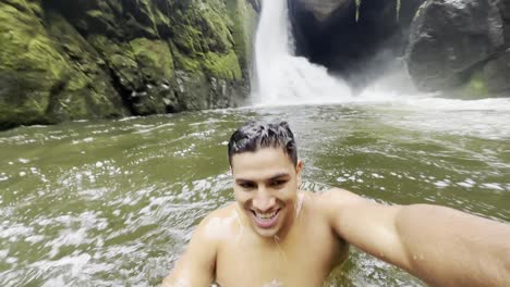 A-young-vlogger-recording-himselft-while-happily-swimming-into-the-lake-near-a-waterfall,-inside-a-rainforest