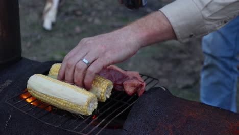 A-close-up-shot-of-a-man-cooking-corn-and-steak-on-a-fire-drum