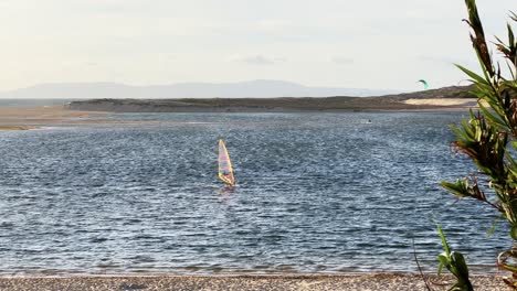 Cinematic-slow-motion-shot-of-a-windsurfer-in-the-foreground-in-Lagoa-de-Albufeira-Portugal,-Europe-and-a-Kitesurfer-in-the-Background-on-a-Sunny-Day-with-a-lot-of-Wind