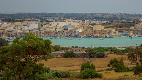 High-angle-shot-over-picturesque-town-along-the-seaside,-off-the-coast-of-Marsaxlokk,-Malta-with-fishing-boats-movement-in-timelapse