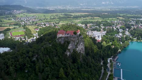 Aerial-drone-shot-of-majestic-castle-on-hilltop-beside-Lake-in-Bled-during-cloudy-day,-Slovenia---Birds-eye-view