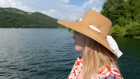 Beautiful-young-blonde-woman-wearing-a-straw-hat-standing-in-front-of-a-lake-and-looking-into-the-distance-with-mountains-and-trees-in-the-back,-profile-view,-sunny-summer-day