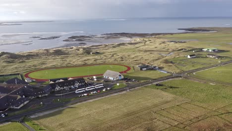 Advancing,-wide-angle-drone-shot-of-the-village-of-Linaclate-in-Benbecula,-featuring-the-Dark-Island-Hotel,-the-local-track-and-football-pitch,-and-the-distant-beach-and-ocean